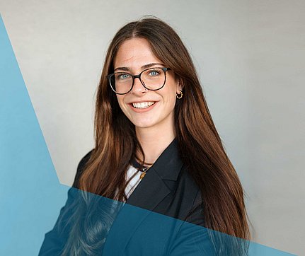 Eine Frau mit langen braunen Haaren und Brille, die lächelt; sie trägt einen schwarzen Blazer mit einem blauen geometrischen Überwurf.