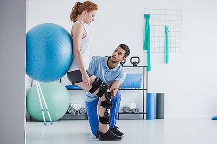 Eine Frau mit Knieorthesen macht in einer Rehabilitationsklinik gemeinsam mit einem Physiotherapeuten Übungen mit einem Gymnastikball.