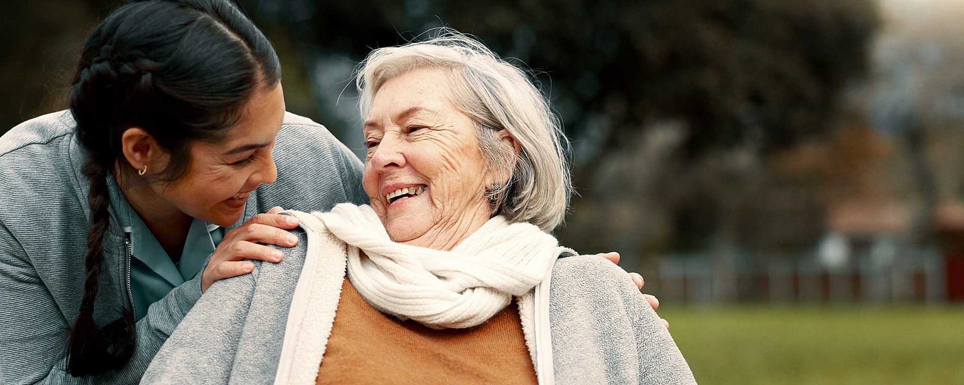 Eine junge Frau und eine ältere Frau lächeln und umarmen sich im Freien und genießen gemeinsam einen fröhlichen und liebevollen Moment.