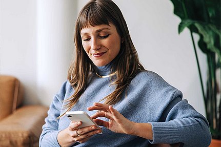 Eine Frau in einem blauen Pullover sitzt drinnen in der Nähe einer Pflanze und lächelt, während sie ihr Smartphone benutzt.