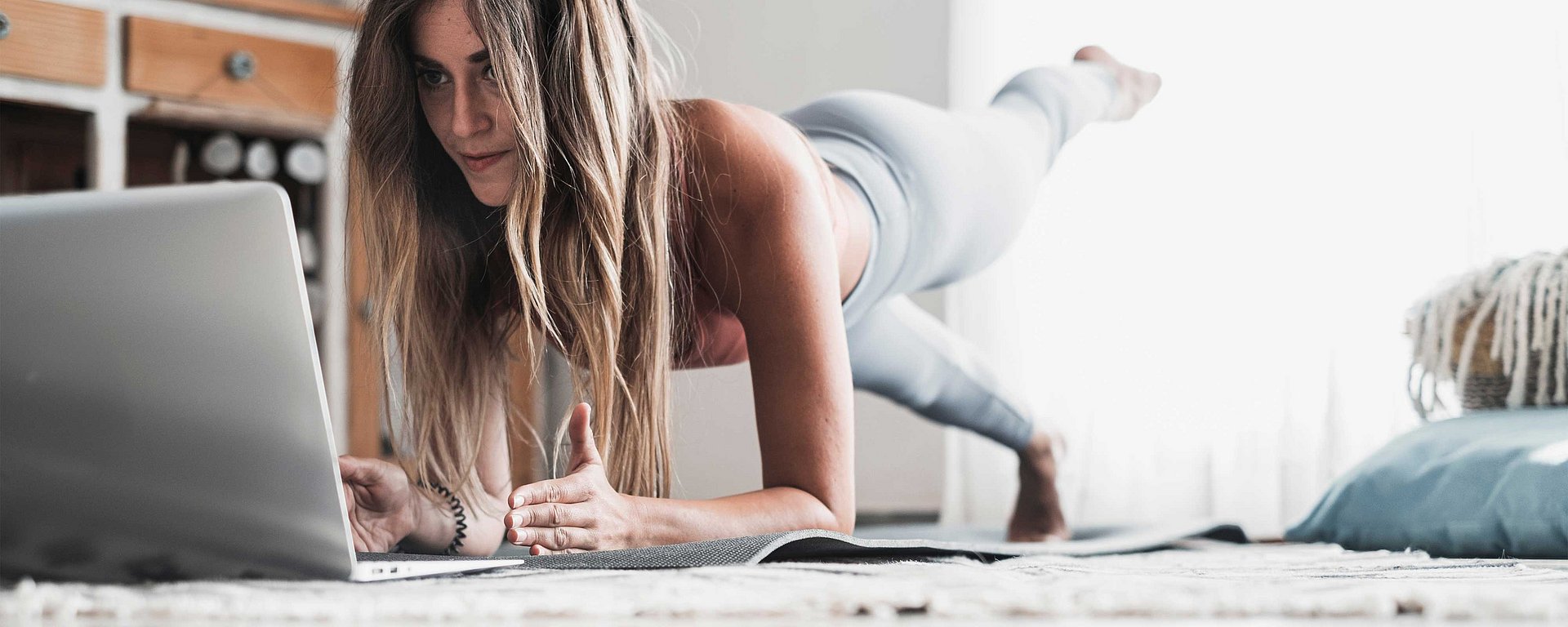 Eine Frau in Sportkleidung, die zu Hause eine Plank-Übung macht, während sie auf einem Laptop etwas anschaut.
