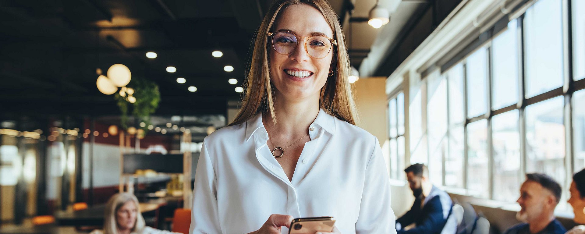 Lächelnde Frau mit Brille, die ein Handy in der Hand hält, in einem modernen Büro, im Hintergrund sind arbeitende Menschen zu sehen.