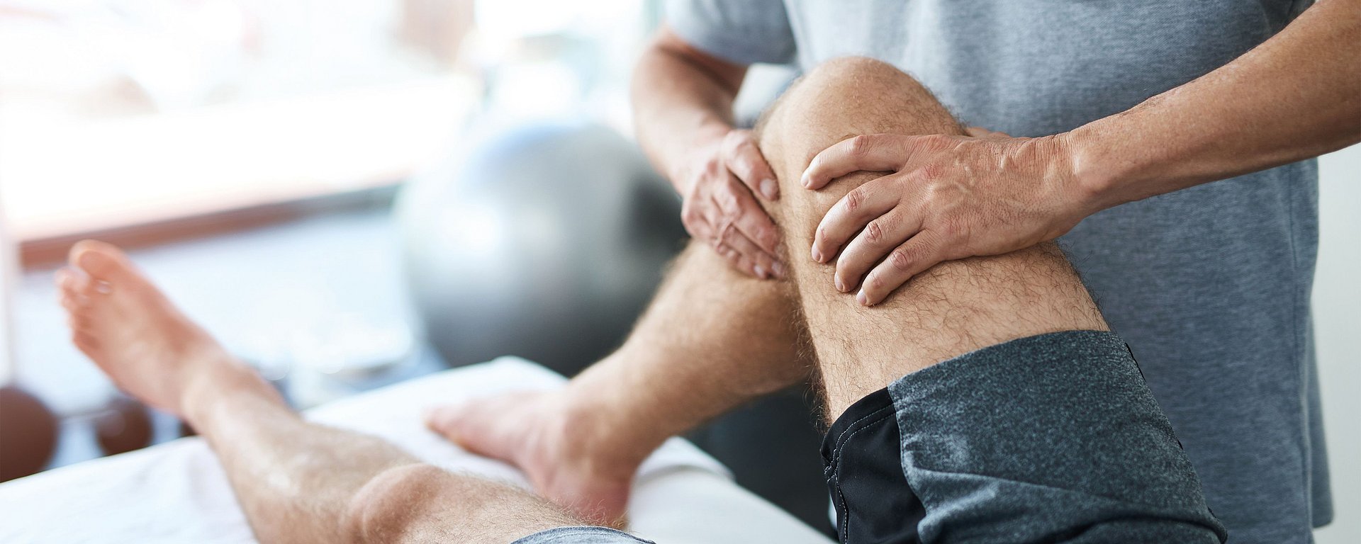 Ein Physiotherapeut untersucht das angewinkelte Knie eines Patienten auf einer Untersuchungsliege in einer Praxis.