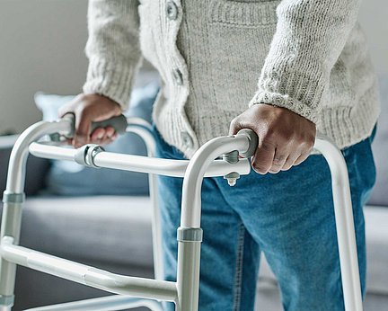 Nahaufnahme einer Person in einem Pullover, die sich in einem Raum mit einem Rollator abstützt.