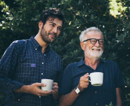 Zwei Männer stehen im Freien, lächeln und halten weiße Kaffeetassen in den Händen, umgeben von viel Grün.